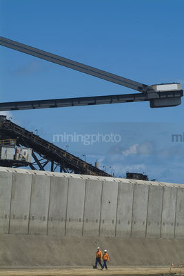 2 mine workers in full PPE  safety gear walk below coal stacker reclaimer at terminal with concrete precast bunding in foreground. vertical image. - Mining Photo Stock Library