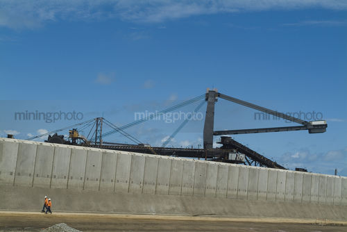 2 mine workers in full PPE  safety gear walk below coal stacker reclaimer at terminal with concrete precast bunding in foreground.  - Mining Photo Stock Library