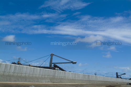 2 mine workers in full PPE  safety gear walk below coal stacker reclaimer at terminal with concrete precast bunding in foreground.  very long panorama shot. - Mining Photo Stock Library