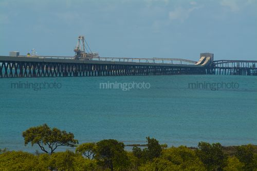 Long jetty out to shipping wharf with dual coal conveyors.  blue ocean and green mangrove foliage in foreground. - Mining Photo Stock Library