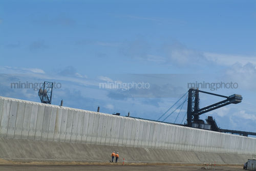 Two mine workers in full ppe walking below pre cast concrete bunding at coal port terminal. reclaimers and ship loaders in the background. great photo depicting scale and would suit panorama - Mining Photo Stock Library