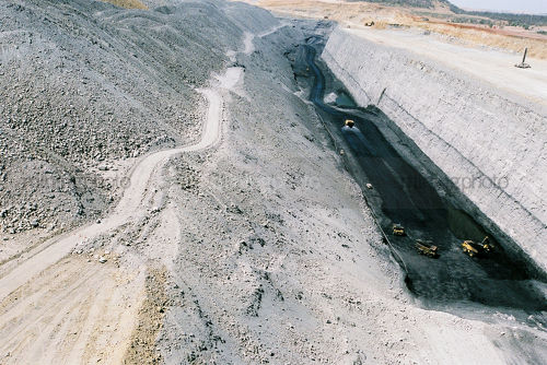 Three truck rotation with digger on coal seam floor with steep high walls.  water cart in background and long haul road behind. midden overburden stockpiles on side.  image shows scale of huge size and depth of mine.   - Mining Photo Stock Library
