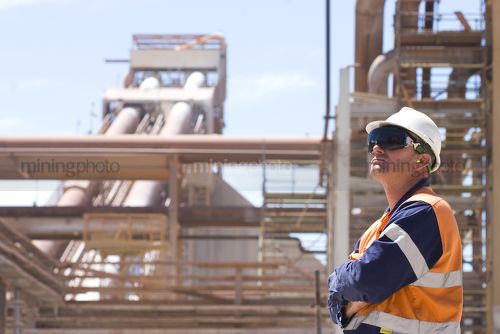 Process engineer onsite in full ppe wearing ear plugs gazing upwards with arms folded.  pipeline and plant in background.  worker is on right hand side of frame.  great generic shot. - Mining Photo Stock Library