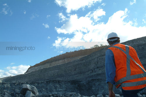 Mine engineer worker inspecting steep high wall from the floor of an open cut coal mine.  shot from behinid and down low.  worker on right hand side of frame. - Mining Photo Stock Library