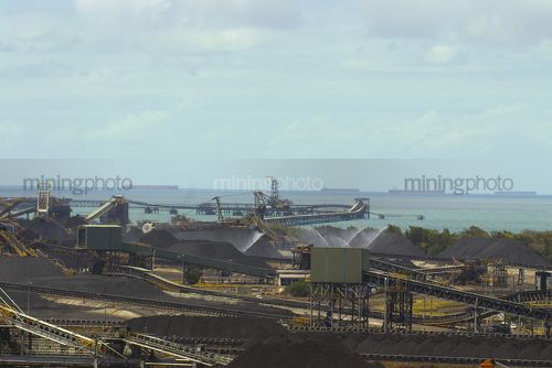 Wide coal terminal shot with stockpiled coal and reclaimers and loaders and lots of ships waiting to be loaded in the background. - Mining Photo Stock Library