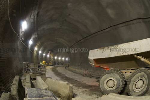 Dump truck inside tunnel works with construction crews on mobile travel tower in background. - Mining Photo Stock Library