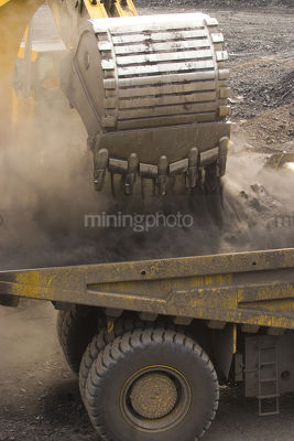 Close up of digger bucket loading haul truck with overburden in open cut mine.  lots of dust. - Mining Photo Stock Library