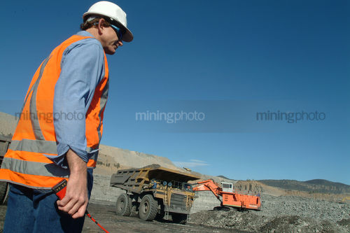 Mine site engineer project manager with hand held radio standing near haul truck and digger in open cut coal mine. - Mining Photo Stock Library