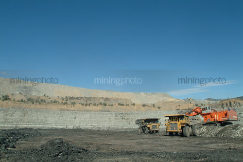Two haul truck rotation with excavator in open cut coal mine.  wide shot with high walls in background. - Mining Photo Stock Library