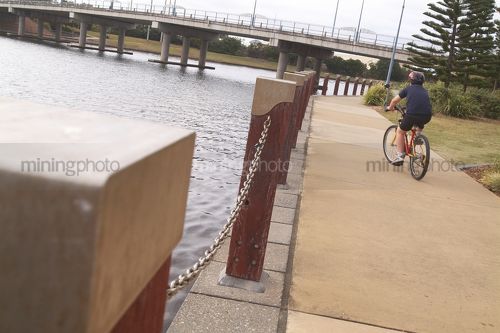 Bike rider along residential waterway with road bridge in background. - Mining Photo Stock Library