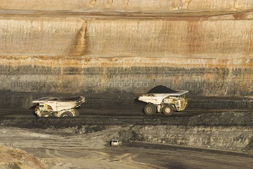 Two haul trucks moving coal.  orange high walls in the afternoon light of an open cut coal mine.  light vehicle in foreground - Mining Photo Stock Library