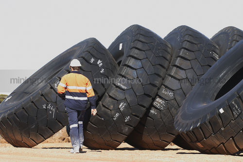 Mine worker in full PPE walking past stockpile of truck tyres.  person is on kleft hand side of frame. plenty of space to the right for copy. - Mining Photo Stock Library