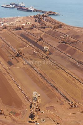 Aerial vertical shot of many reclaimers working and loading iron ore onto conveyors.  ship and shipping port terminal in background with ocean. - Mining Photo Stock Library