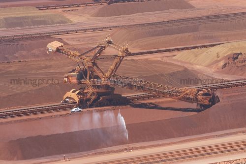 Iron ore reclaimer working at ship terminal. water sprayer for dust suppression with light vehicle adjacent for scale. - Mining Photo Stock Library