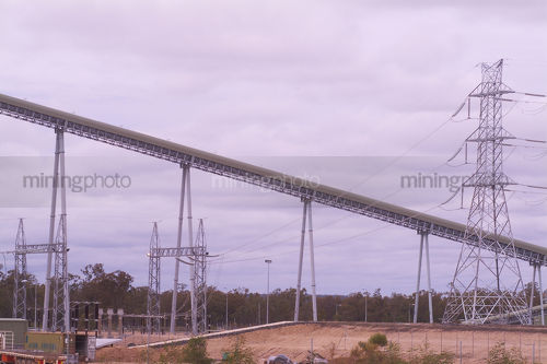 Coal conveyor rising high at power station with transmission tower in foreground. - Mining Photo Stock Library