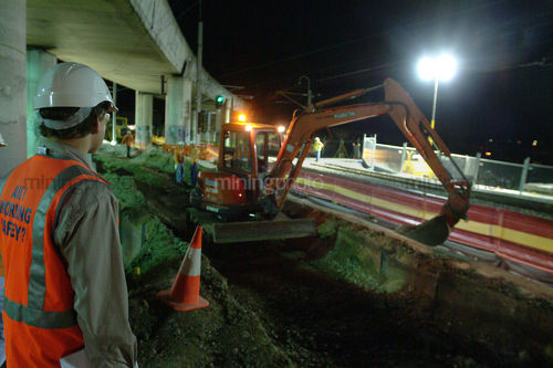 Project manager in full PPE overseeing night works on train line with excavator.  shto at night with construction lighting. - Mining Photo Stock Library