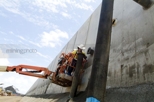 Worker in ppe operating cherry picker adjacent to huge concrete precasts with coal loaders in the background - Mining Photo Stock Library