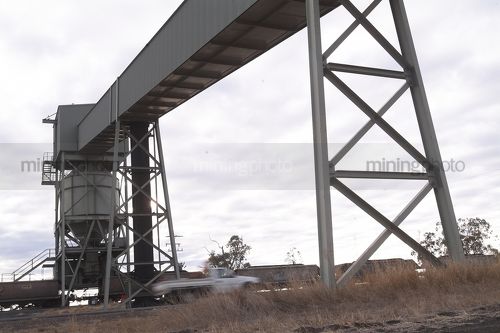 Farmer in light vehicle ute passes under overhead coal conveyor loading rail trains in remote, rural area.  car is moving. - Mining Photo Stock Library