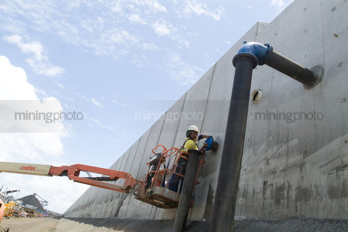 Worker in harness and full PPE fits cover to water pip from cherry picker bucket.  this is adjacent to large pre cast concrete bunding - Mining Photo Stock Library