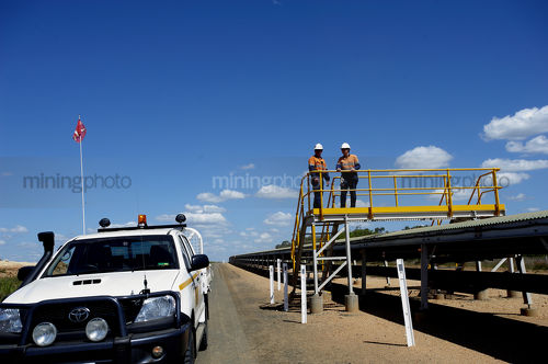 Two mine workers in full PPE standing on pedestrian overpass with overland covered coal conveyor below. conveyor stretches into background and light vehicle on access road adjacent. - Mining Photo Stock Library