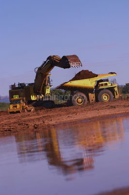 Excavator loading overburden into haul truck in late afternoon light.  reflections on water in foreground. - Mining Photo Stock Library