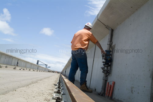 Worker in PPE testing a valve on coal loader track at ship terminal. shot from behind - Mining Photo Stock Library
