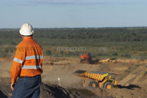 Mine worker in full PPE observing haul truck and mine workings at open cut mine. - Mining Photo Stock Library