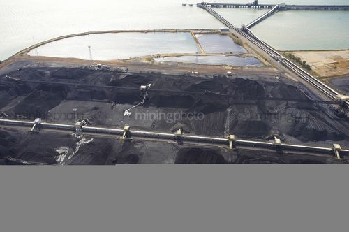Aerial photo of stockpiled coal at shipping terminal with conveyors, loaders and large ship wharf in background. - Mining Photo Stock Library