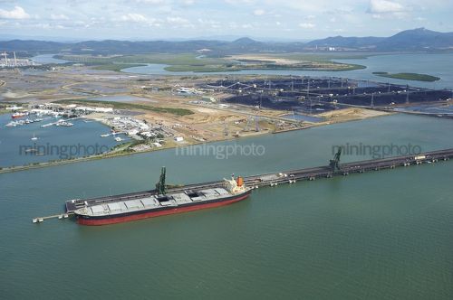 Wide aerial shot of a coal ship being loaded at wharf.  stock piled coal in background with reclaimers.  marina close by. - Mining Photo Stock Library