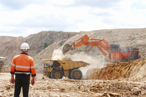 Male worker in full PPE observing haul truck being loaded with overburden by excavator in open cut mine. - Mining Photo Stock Library