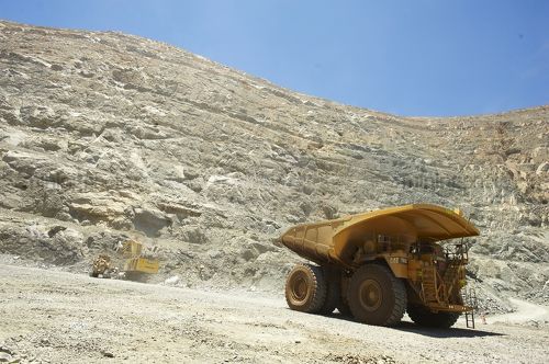 Wide shot at ground level of loaded haul truck leaving the deep open cut pit floor.  excavator and dozer in background at base of high wall.  blue sky and high walls above.  gold mine. - Mining Photo Stock Library