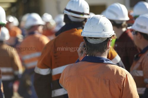 Mine worker in full PPE at site meeting with lots of workers in background.  very generic shot with no faces and those in the background out of focus. - Mining Photo Stock Library