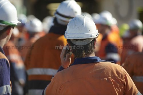 Mine worker in full PPE at site meeting with lots of workers in background.  very generic shot with no faces and those in the background out of focus. - Mining Photo Stock Library