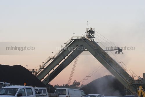 Coal stockpile conveyor with walkway on mine site inearly morning light.  light vehicles parked up in site carpark in foreground. - Mining Photo Stock Library