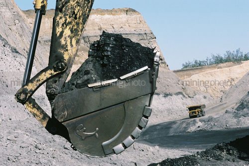 Excavator bucket full of coal in foreground with haul truck returning down haul access road in background.  high walls of coal seam and open cut mine. - Mining Photo Stock Library
