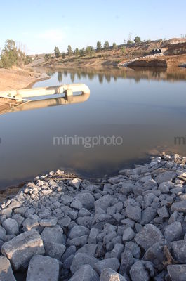 Pumping pipe in water storage dam on a mine site.  drainage rocks in foreground. vertical image. - Mining Photo Stock Library