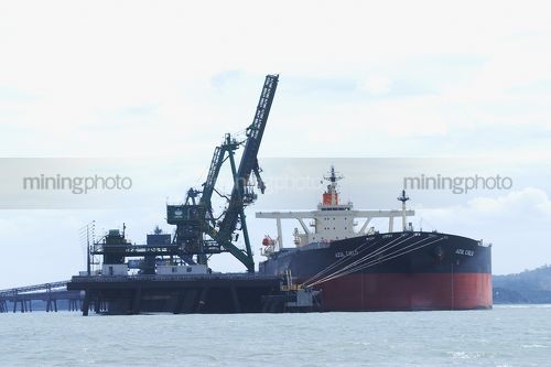 Ship berthed at coal wharf and being loaded by ship loader.  photo taken from a boat at water level. - Mining Photo Stock Library