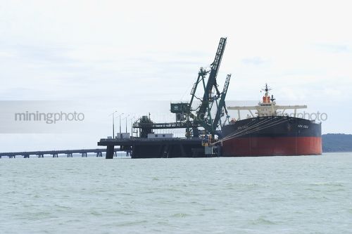 Ship berthed at coal wharf and being loaded by ship loader.  photo taken from a boat at water level. - Mining Photo Stock Library