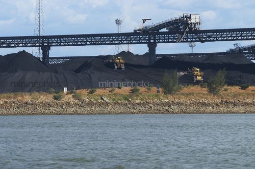 Two dozers stock piling coal at wharf terminal.  overhead conveyor in background.  sea water in foreground. - Mining Photo Stock Library
