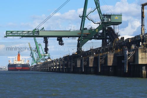 Close up photo of ship loader loading coal into ship at wharf.  shot from water level behind the ship. - Mining Photo Stock Library