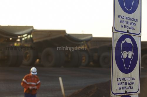 Hearing and head protection safety signs in focus with mine haul truck driver in full PPE walking from truck go line in background. - Mining Photo Stock Library