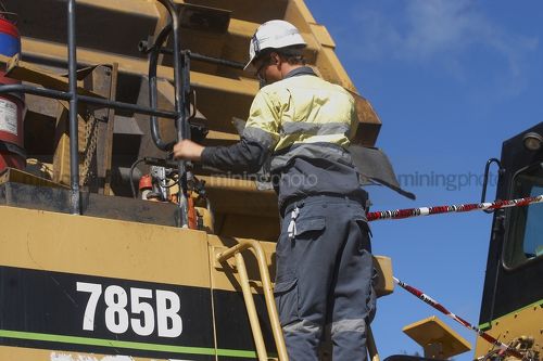 Mine worker in full PPE including harness climbs down ladder on the front of a haul truck he has been maintaining. - Mining Photo Stock Library