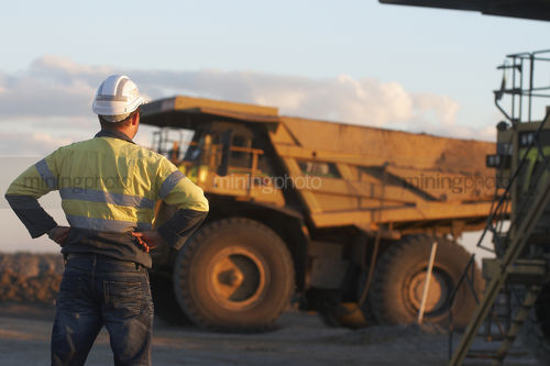 Worker in full PPE with hands on hips observing haul trucks at the go line.  shot in the late afternoon light. - Mining Photo Stock Library