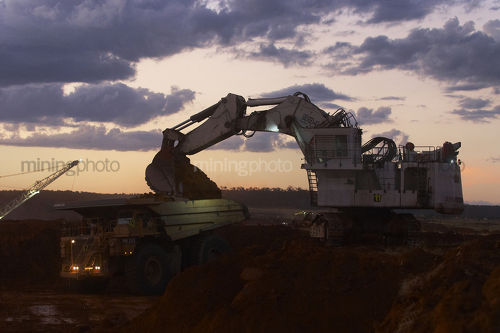 Excavator loading overburden into a haul truck in an open cut coal mine.  late afternoon dusk image. - Mining Photo Stock Library
