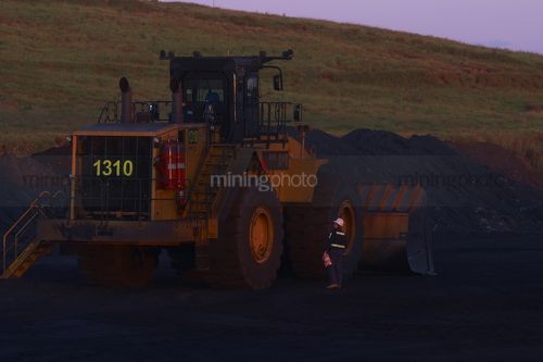 Worker in full PPE inspecting loader in open cut mine workshop area.  shot in late afternoon dusk light. - Mining Photo Stock Library