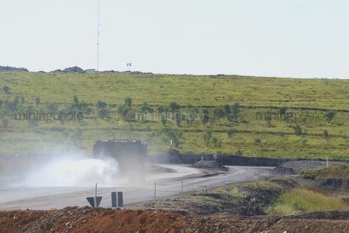 Water cart spraying water on haul access road near workshop with mine site revegetation on hill in background. - Mining Photo Stock Library