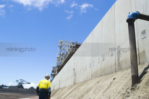 Mine worker walking along bunding at coal terminal.  coal reclaimers in the background. - Mining Photo Stock Library
