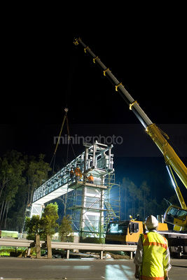 Civil worker supervising gantry being lifted into palce by crane.  workers on scaffold fitting gantry into place.  night time works. - Mining Photo Stock Library