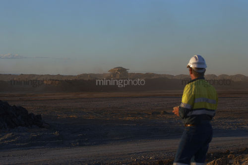 Mine site worker in full PPE observing haul truck in late afernoon light in open cut mining site. - Mining Photo Stock Library