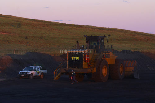 Mine site worker walking around parked loader at the end of a shift.  light vehicle parked adjacent. shot in late afternoon dusk light. - Mining Photo Stock Library